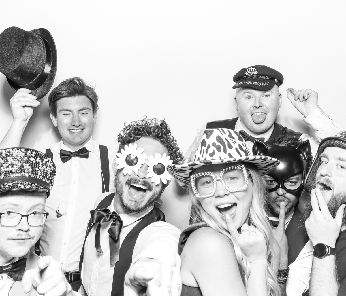 large group of wedding guests posing against a plain white backdrop, during the party entertainment in the evening, wearing fun party props and laughing for the camera, with black and white prints for the kardashian style photos
