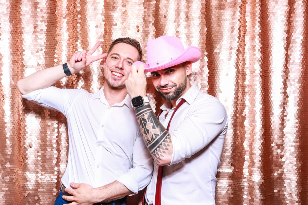 2 guests posing against a blush sequins backdrop, for an open photo booth for weddings, during the party entertainment in the evening, wearing fun party props and laughing for the camera