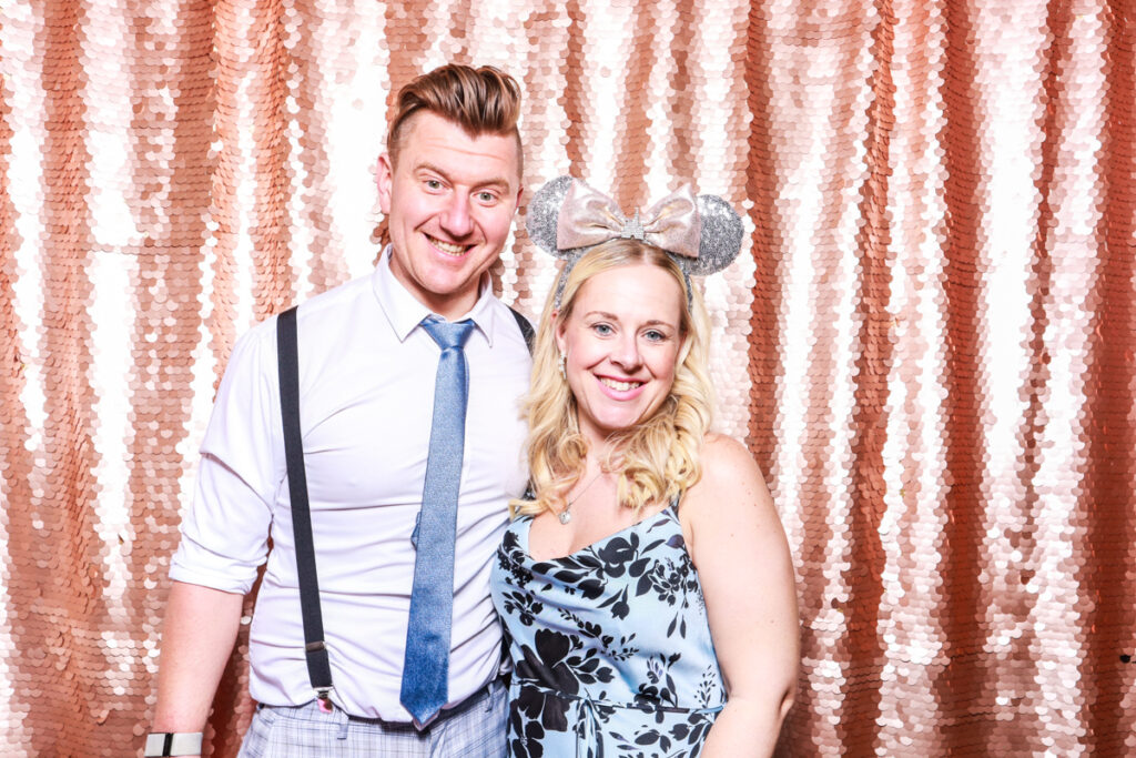 2 guests posing against a blush sequins backdrop, for an open photo booth for weddings, during the party entertainment in the evening, wearing fun party props and laughing for the camera