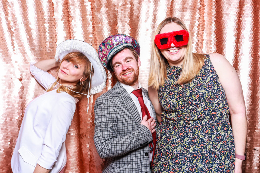 3 guests posing against a blush sequins backdrop, for an open photo booth for weddings, during the party entertainment in the evening, wearing fun party props and laughing for the camera