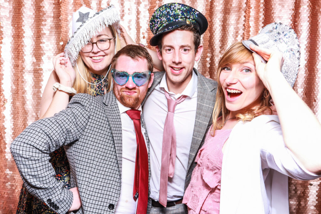4 guests posing against a blush sequins backdrop, for an open photo booth for weddings, during the party entertainment in the evening, wearing fun party props and laughing for the camera