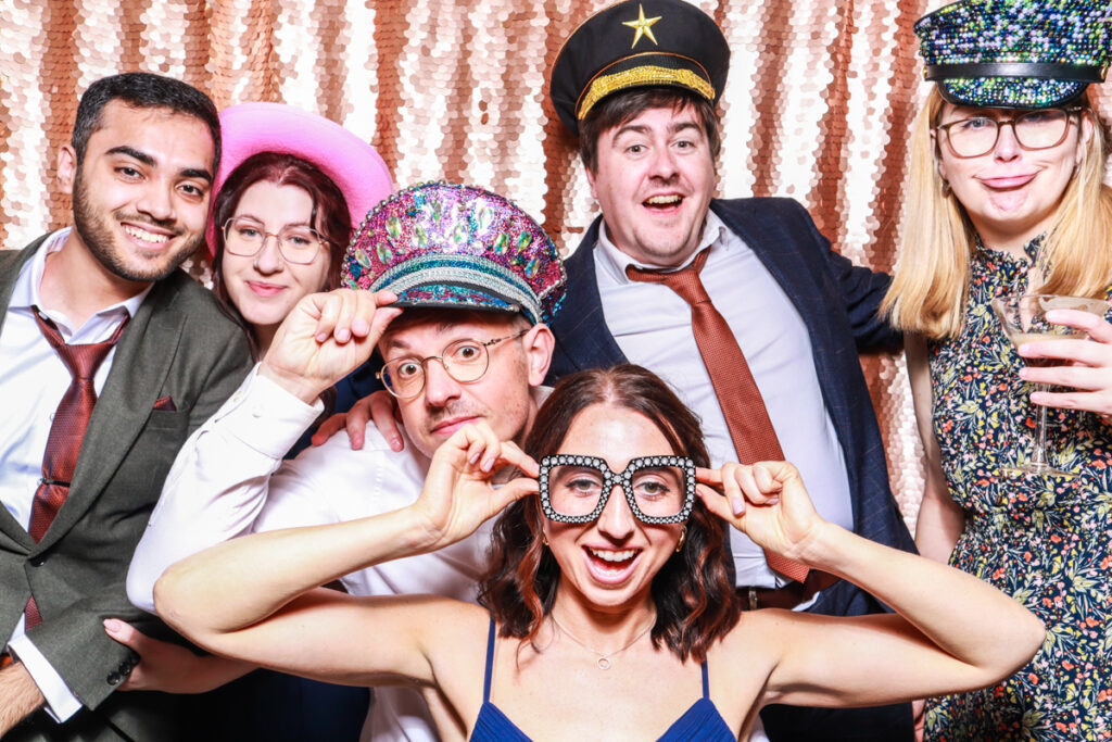 large group of wedding guests posing against a blush sequins backdrop, for an open photo booth for weddings, during the party entertainment in the evening, wearing fun party props and laughing for the camera