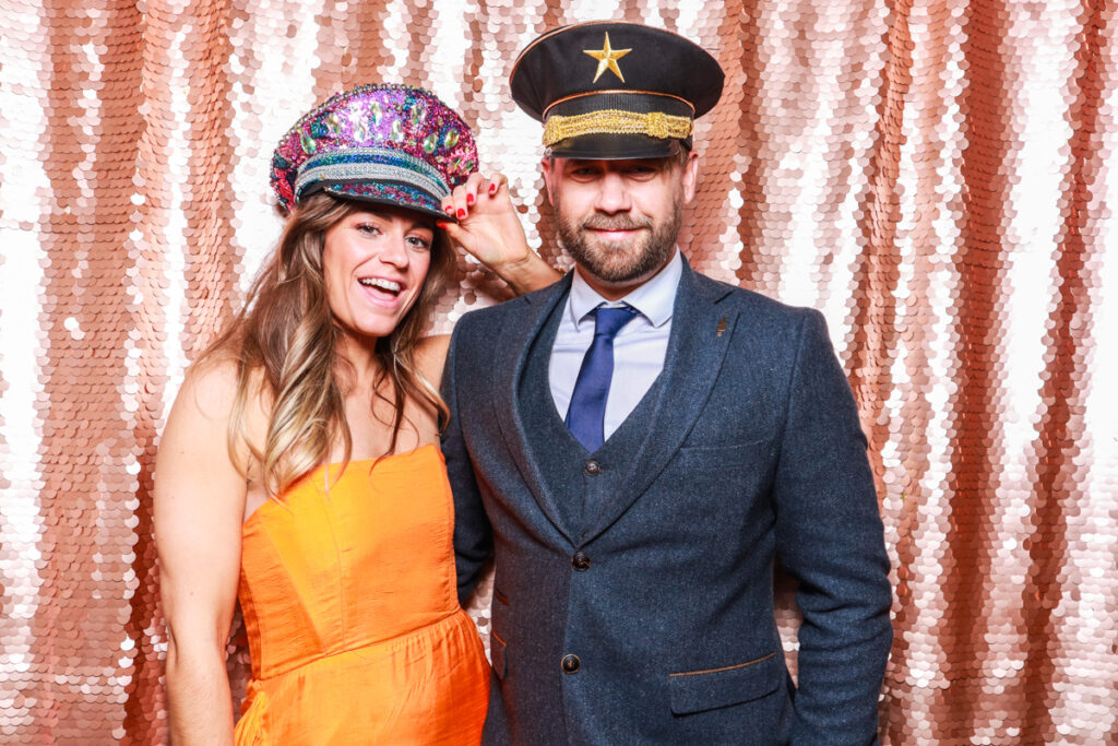 2 guests posing against a blush sequins backdrop, for an open photo booth for weddings, during the party entertainment in the evening, wearing fun party props and laughing for the camera