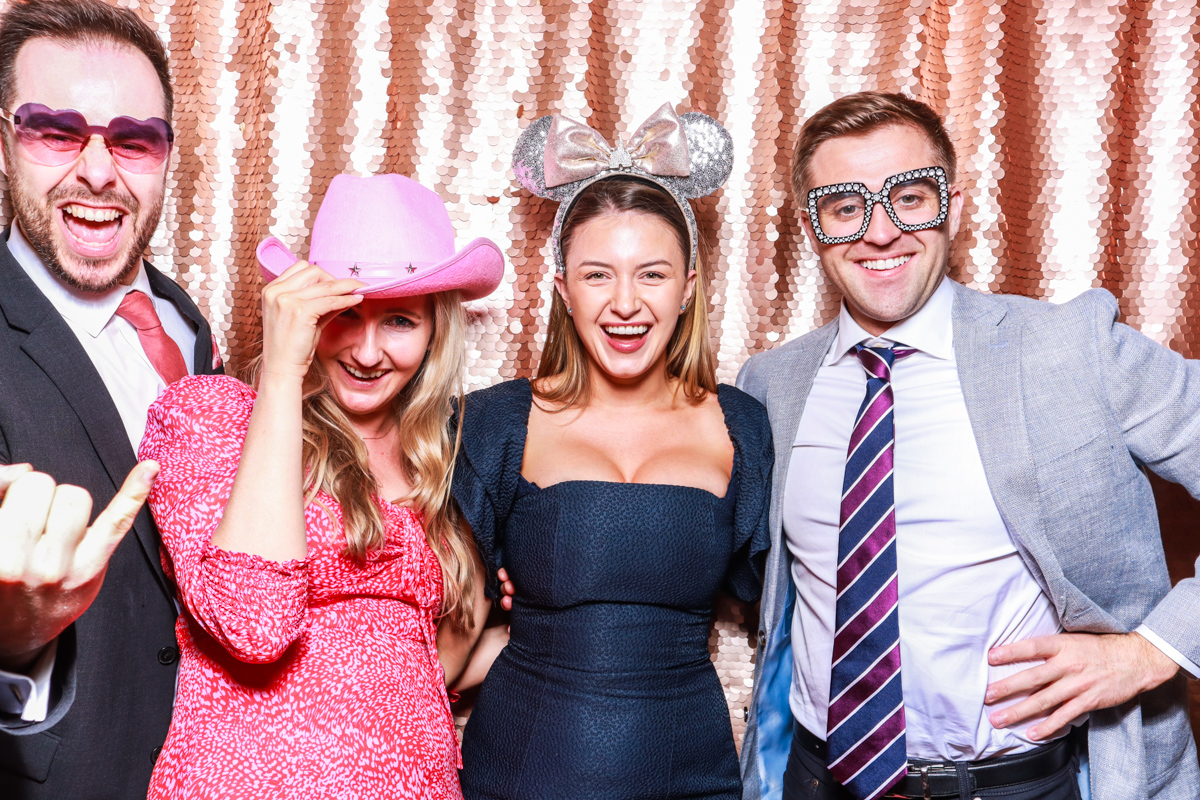 4 guests posing against a blush sequins backdrop, for an open photo booth for weddings, during the party entertainment in the evening, wearing fun party props and laughing for the camera