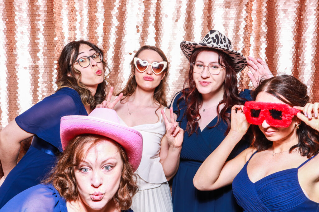 5 guests posing against a blush sequins backdrop, for an open photo booth for weddings, during the party entertainment in the evening, wearing fun party props and laughing for the camera