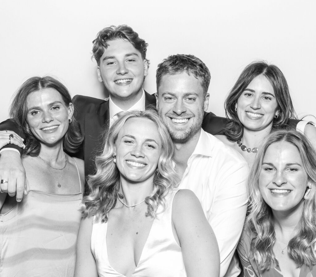 large group of wedding guests posing against a plain white backdrop, during the party entertainment in the evening, wearing fun party props and laughing for the camera, with black and white prints for the kardashian style photos