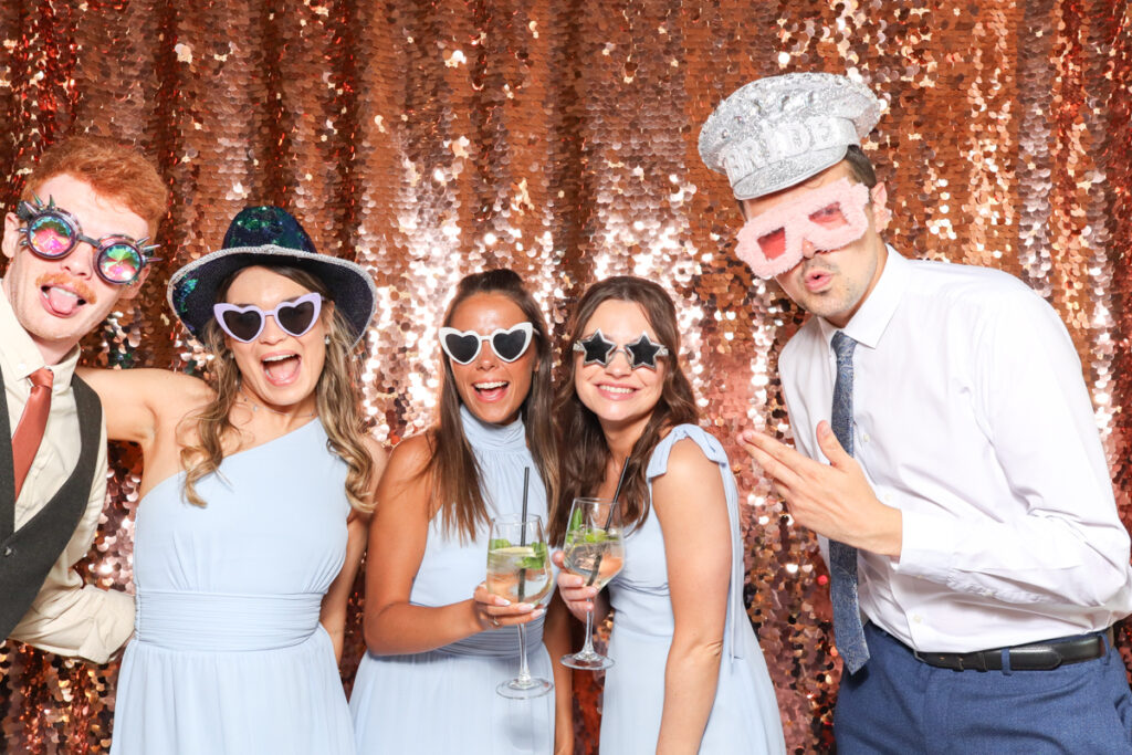 5 guests posing against a champagne sequins backdrop, for an open photo booth for weddings, during the party entertainment in the evening, wearing fun party props and laughing for the camera