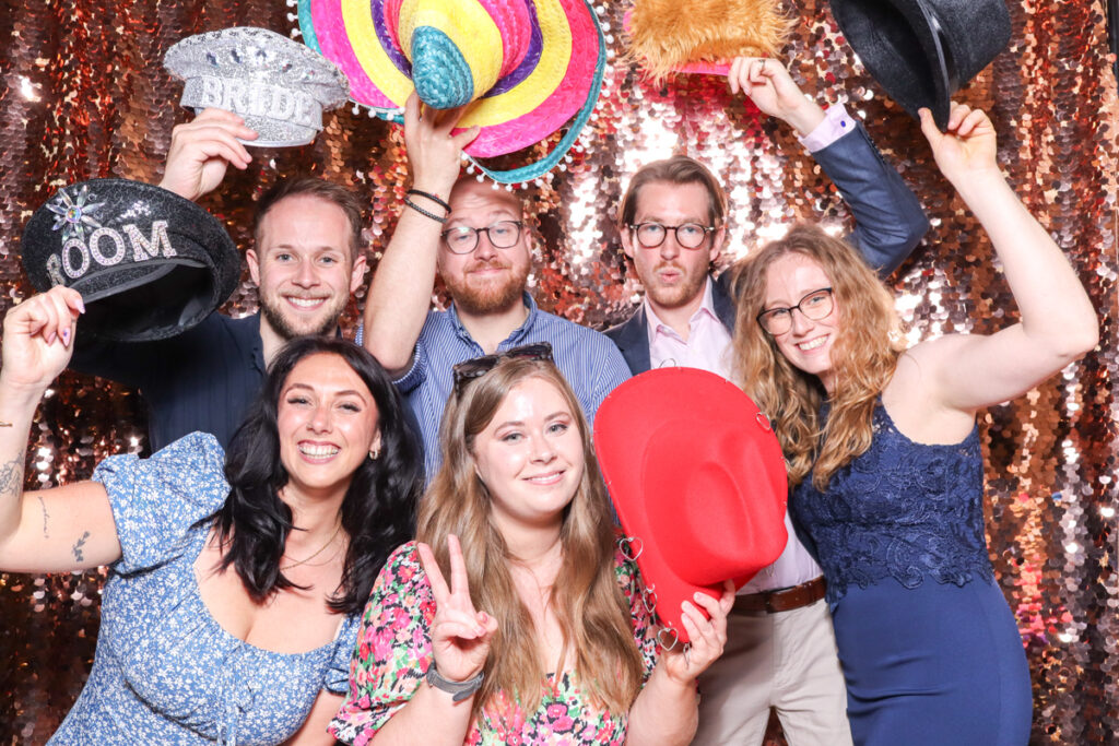 large group of wedding guests posing against a champagne sequins backdrop, for an open photo booth for weddings, during the party entertainment in the evening, wearing fun party props and laughing for the camera