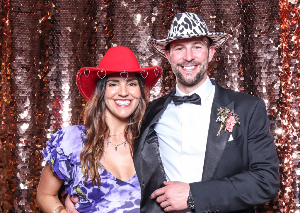 2 smartly dressed guests posing against a champagne sequins backdrop, for an open photo booth for weddings, during the party entertainment in the evening, wearing fun party props and laughing for the camera