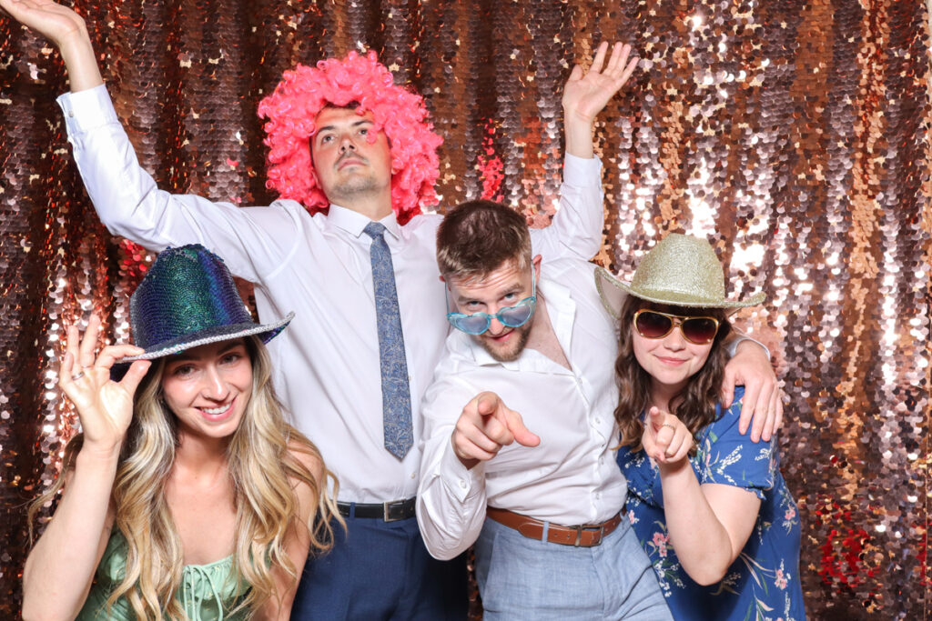 wedding guests posing against a champagne sequins backdrop, for an open photo booth for weddings, during the party entertainment in the evening, wearing fun party props and laughing for the camera