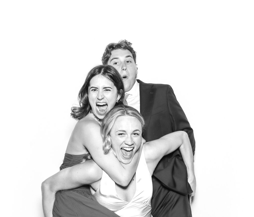 3 wedding guests posing against a plain white backdrop, during the party entertainment in the evening, wearing fun party props and laughing for the camera, with black and white prints for the kardashian style photos