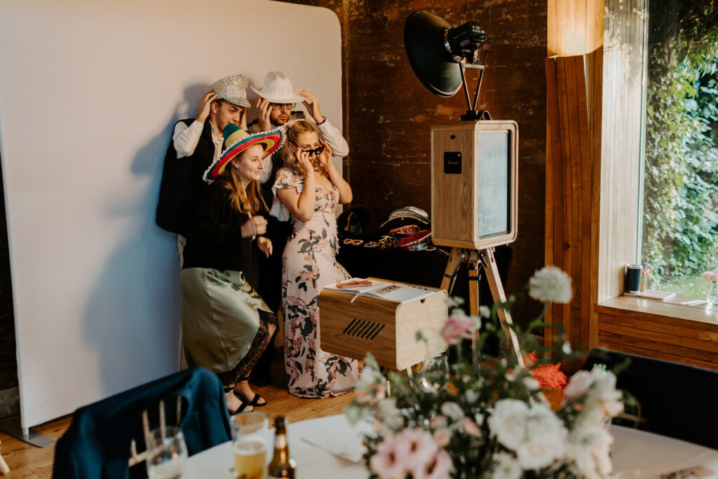 setup of the icon wooden photo booth setup with guests posing wearing party props, posing against a white backdrop for the kardashian style b&w prints