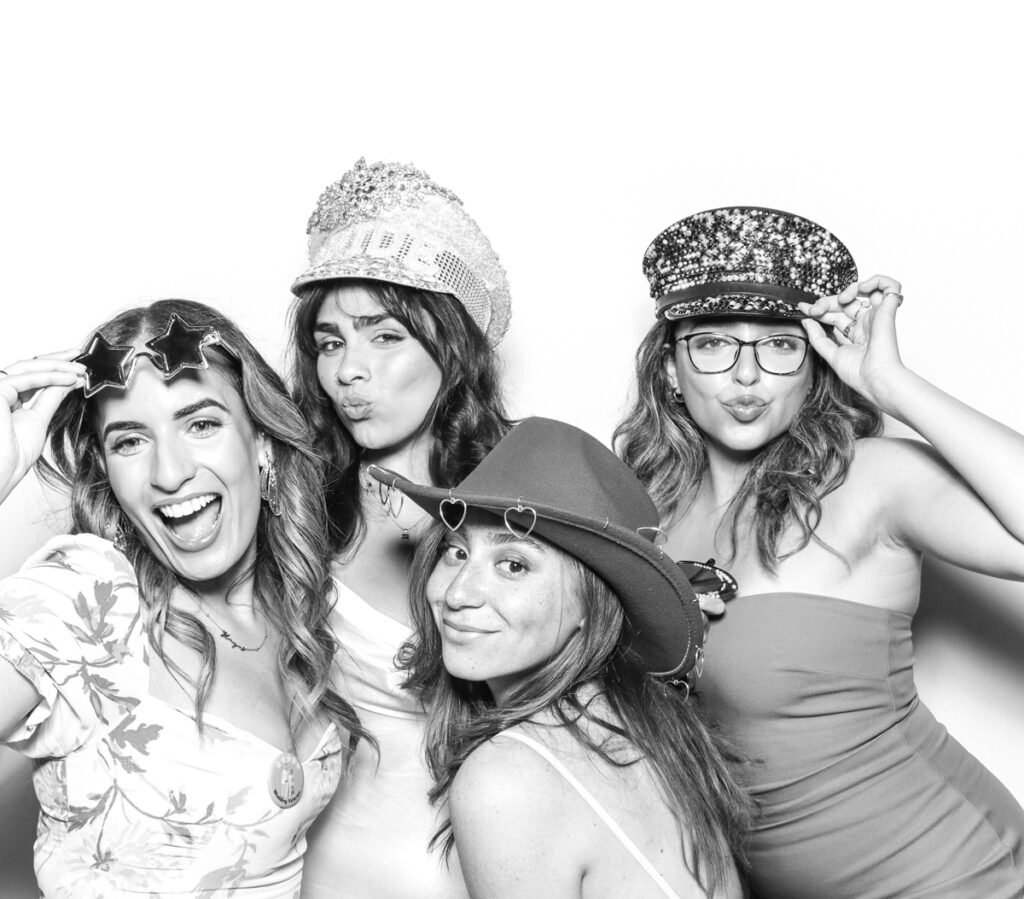 4 wedding guests posing against a plain white backdrop, during the party entertainment in the evening, wearing fun party props and laughing for the camera, with black and white prints for the kardashian style photos