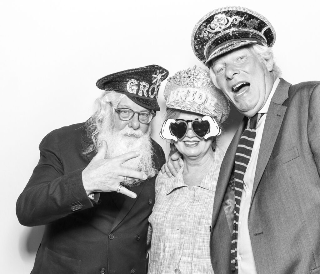3 wedding guests posing against a plain white backdrop, during the party entertainment in the evening, wearing fun party props and laughing for the camera, with black and white prints for the kardashian style photos
