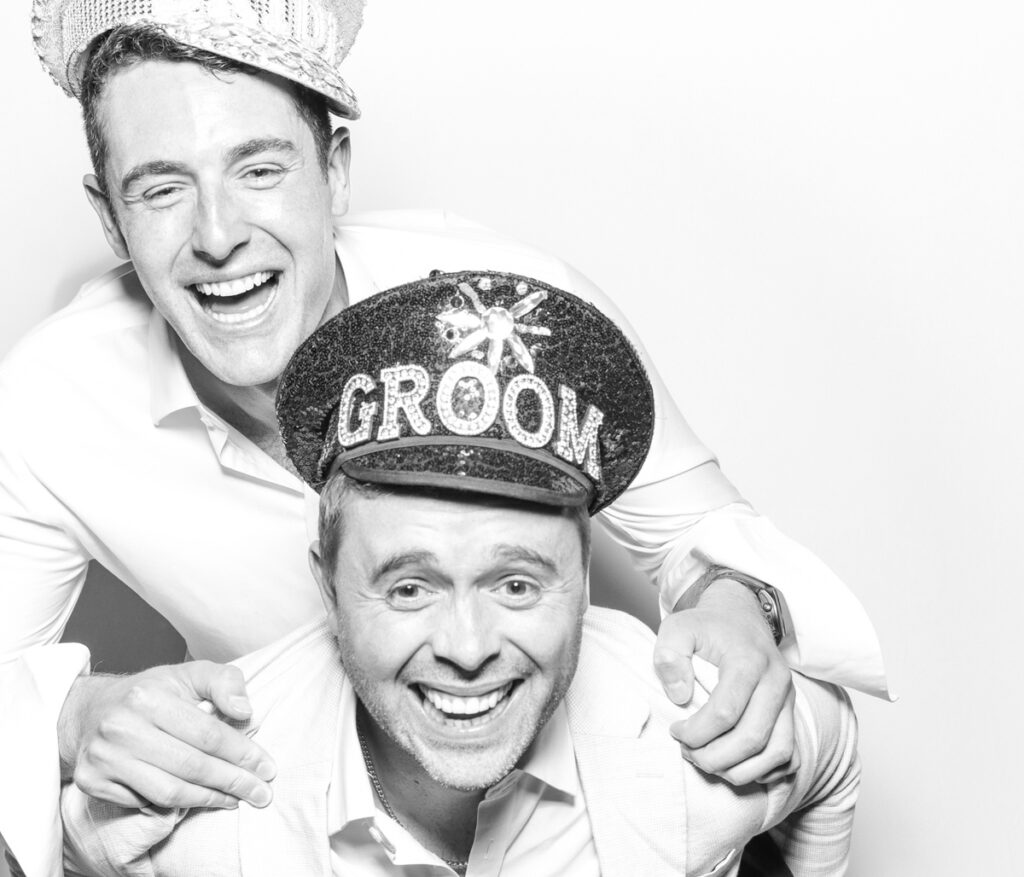 2 wedding guests posing against a plain white backdrop, during the party entertainment in the evening, wearing fun party props and laughing for the camera, with black and white prints for the kardashian style photos