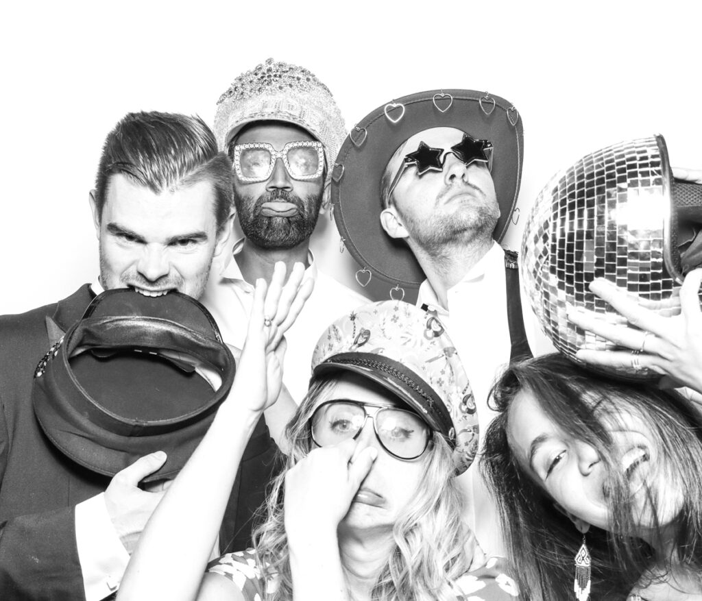 large group of wedding guests posing against a plain white backdrop, during the party entertainment in the evening, wearing fun party props and laughing for the camera, with black and white prints for the kardashian style photos