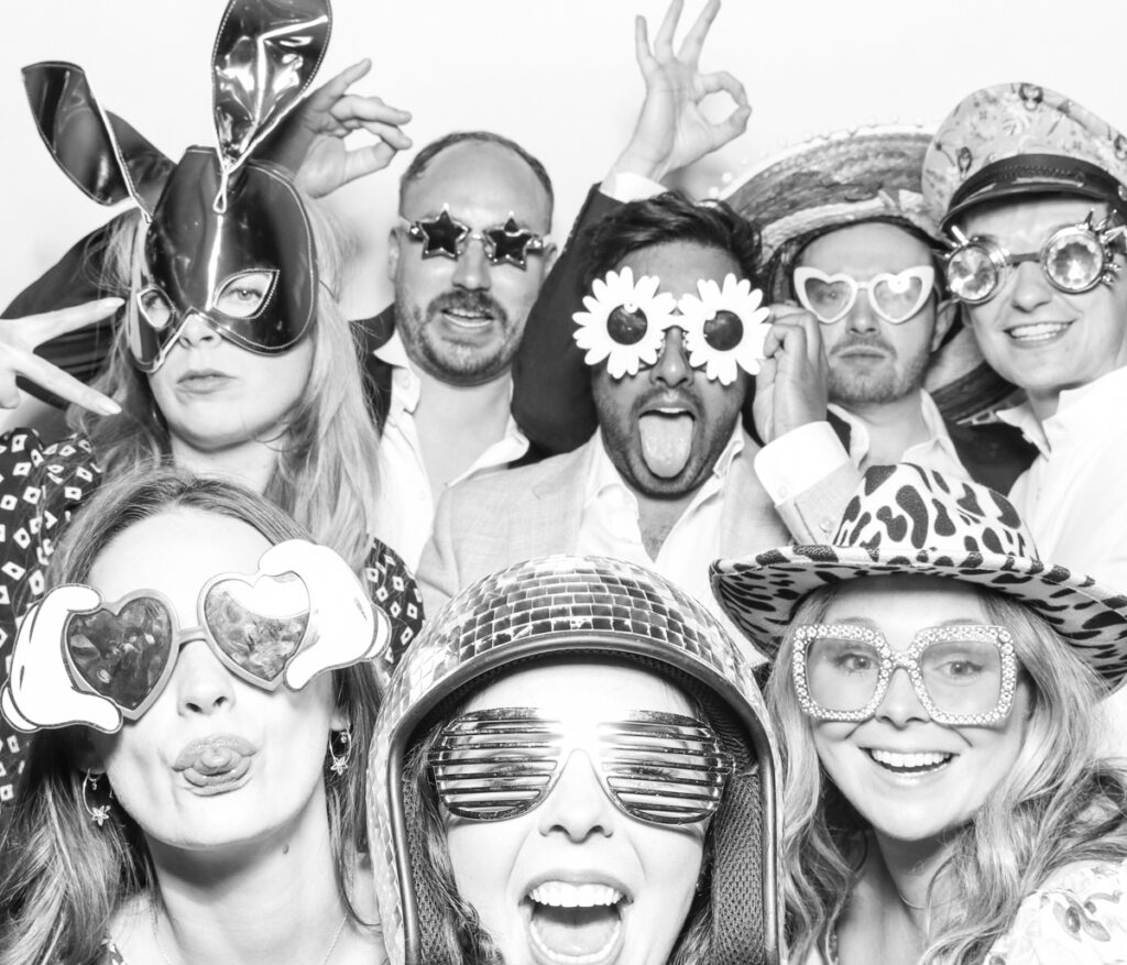 large group of wedding guests posing against a plain white backdrop, during the party entertainment in the evening, wearing fun party props and laughing for the camera, with black and white prints for the kardashian style photos