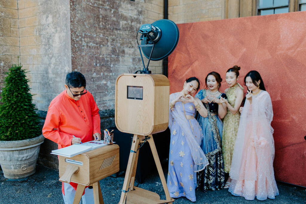 asian guests dressed up posing for a wooden outdoor photo booth with maroon backdrop during an outdoor asian wedding in the summer at grittleton house near Bath