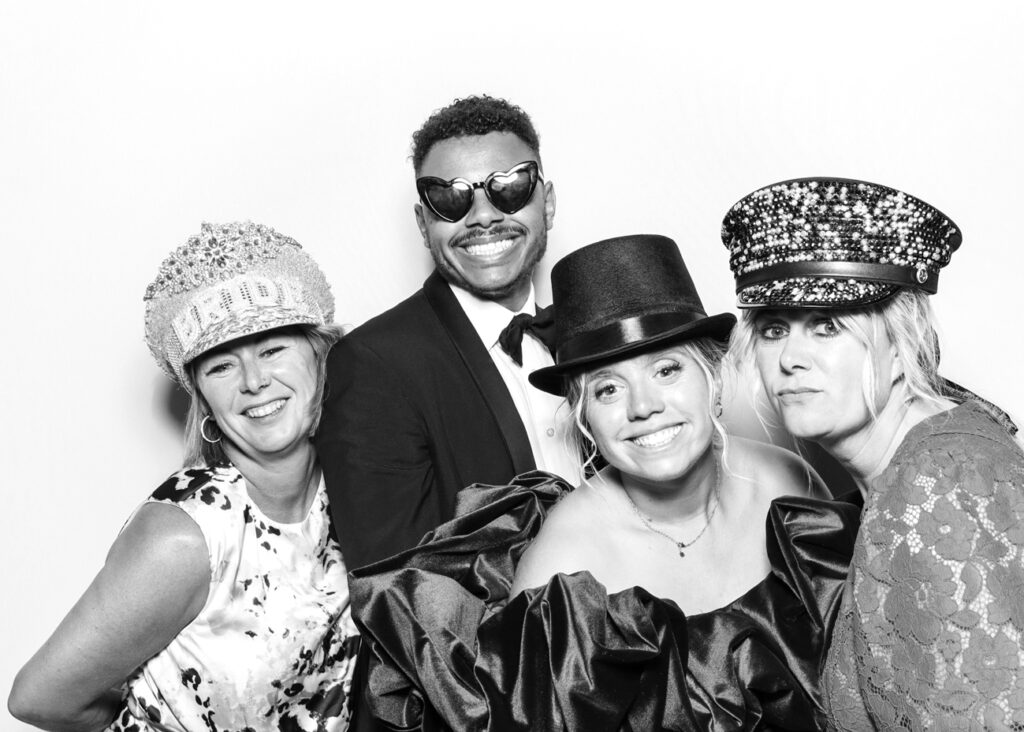 Four wedding guests smile and pose in top hats, heart-shaped sunglasses, and sequinned “Bride” caps in a crisp black & white photo for an Eastnor Castle wedding