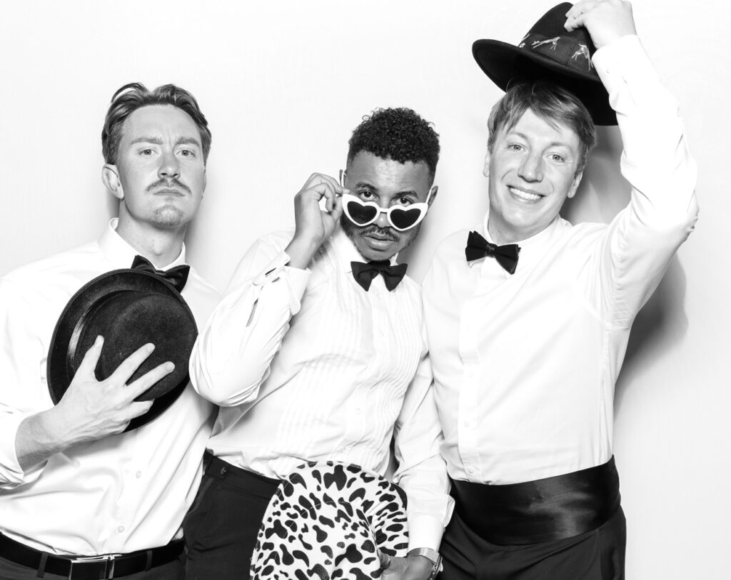 Elegant black and white booth portrait of three men in tuxedos and playful hats during a celebration at Eastnor Castle.