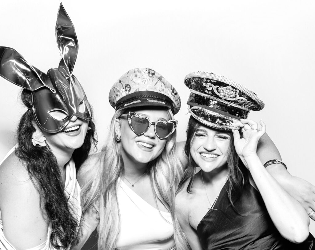 Three guests laughing and enjoying the booth experience with playful hats and accessories in crisp black & white style.