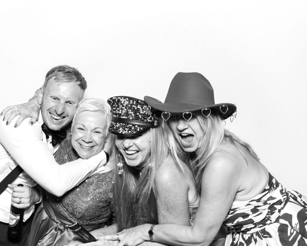 Group of four guests laughing and embracing, wearing sparkly and heart-themed hats in a fun black and white photo booth shot.