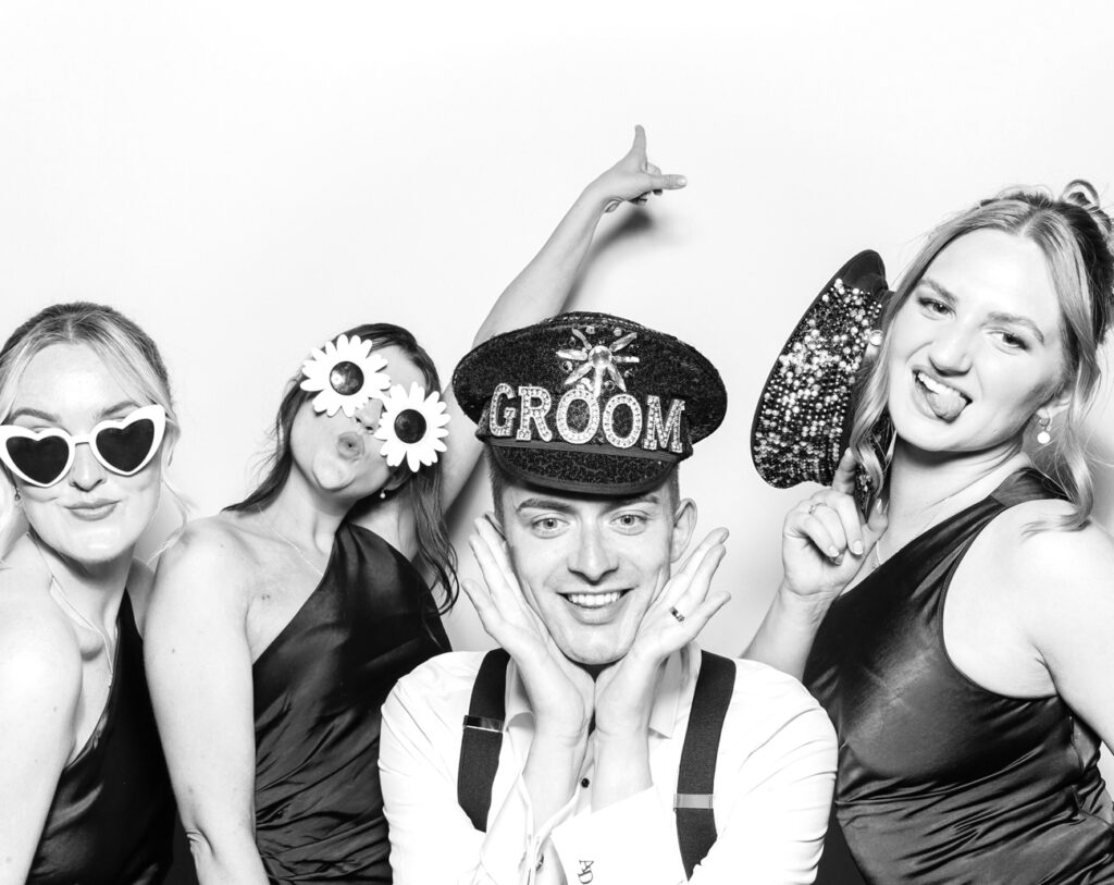 The groom and three guests pose with oversized glasses, glitter hats, and cheeky expressions in a stylish monochrome booth photo for the Eastnor castle wedding
