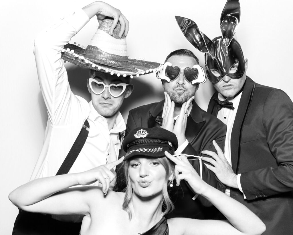 Three men in suits and a woman in formalwear wear sombreros, heart glasses, and bunny masks for a bold black & white booth moment.