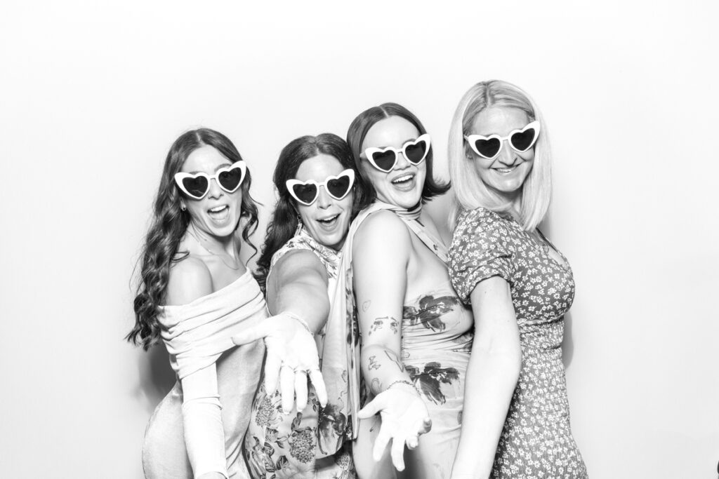 black and white image of 4 guests posing with white heart-shaped sunglasses for photo booths for hire with a kardashian style look , for mad hat photo booth in the cotswolds