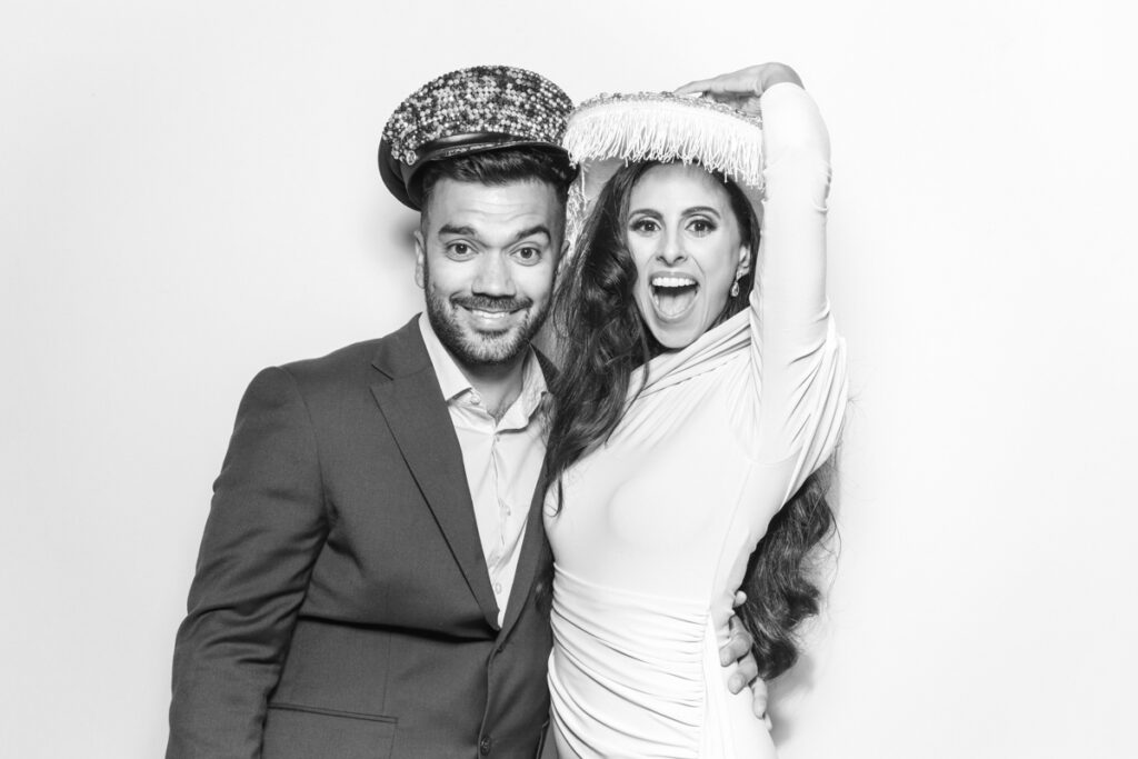 black and white image of 2 guests posing against a white backdrop wearing 2 fun party hats, for a photo booth hire near me with a kardashian style look , for a cotswolds party entertainment