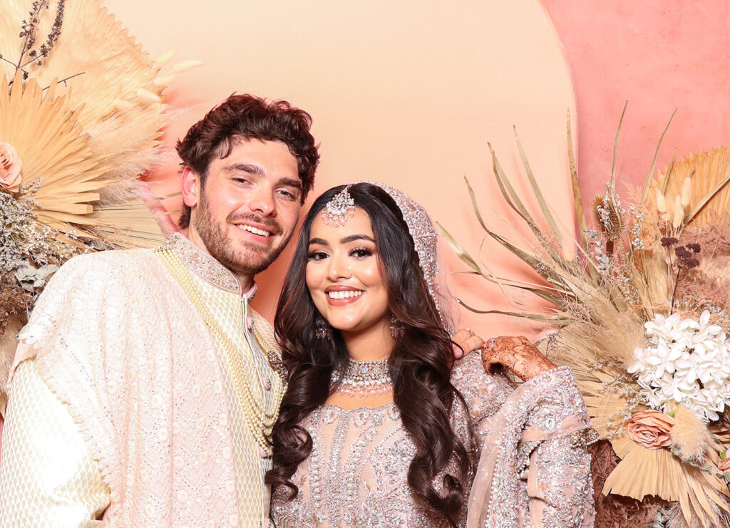 Bride and groom smiling in front of a luxury floral backdrop, dressed in coordinated pastel South Asian wedding outfits with intricate embroidery and soft glam styling
