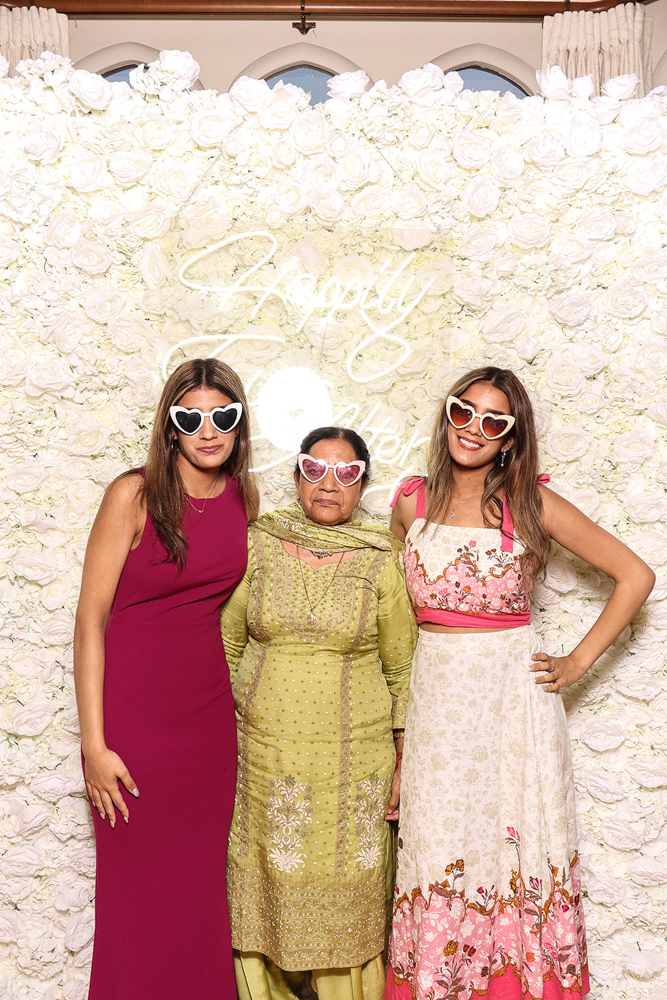 Three women posing in front of a white rose flower wall, wearing heart-shaped sunglasses and colourful South Asian outfits
