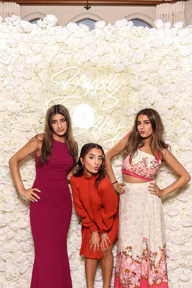 Three women striking playful poses in front of a floral wall with 'Happily Ever After' neon sign, wearing vibrant dresses