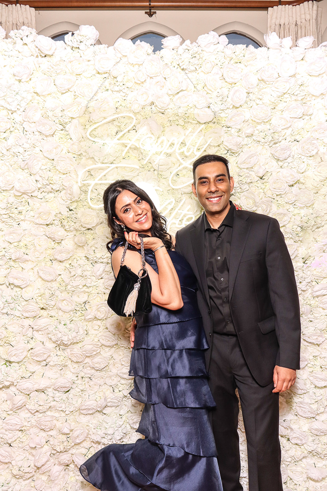 Elegant couple in formalwear smiling in front of a luxury white rose backdrop at a wedding photo booth