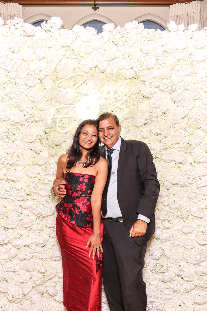 Couple dressed in classic wedding guest attire posing with smiles in front of a white rose wall
