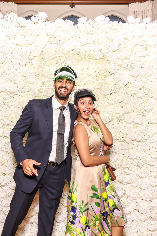 Playful couple with floral dress and suit, both wearing visors and posing excitedly in front of a rose backdrop
