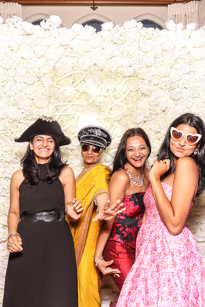 Group of four women wearing hats, sunglasses and bright dresses, having fun in front of a flower wall at a wedding celebration