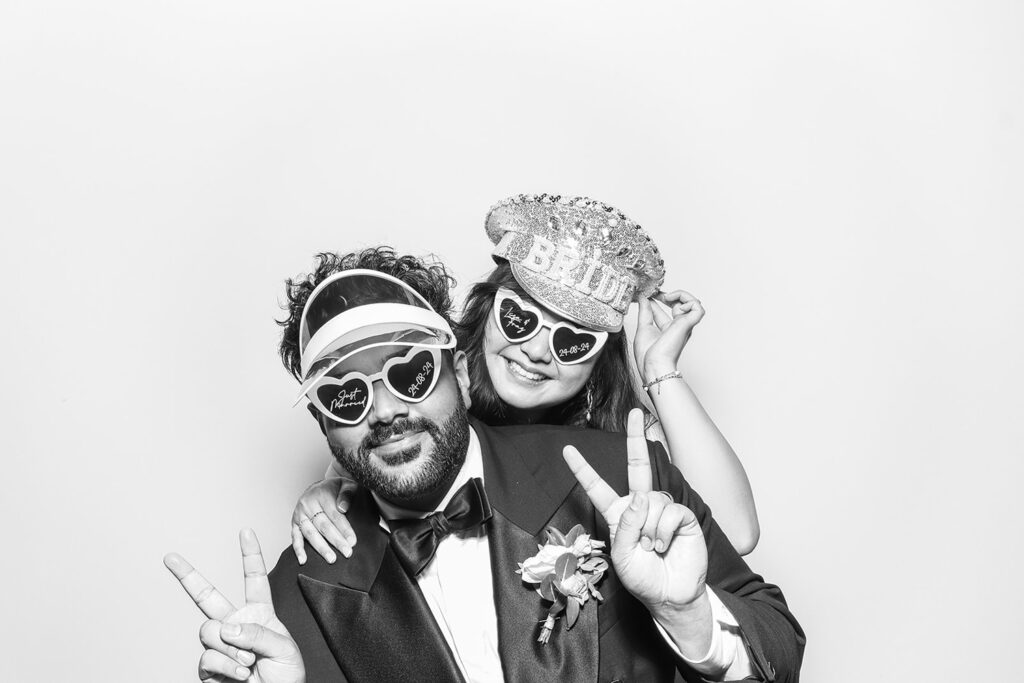 Bride and groom posing with heart-shaped glasses and peace signs in a Kardashian-style black and white photo booth print