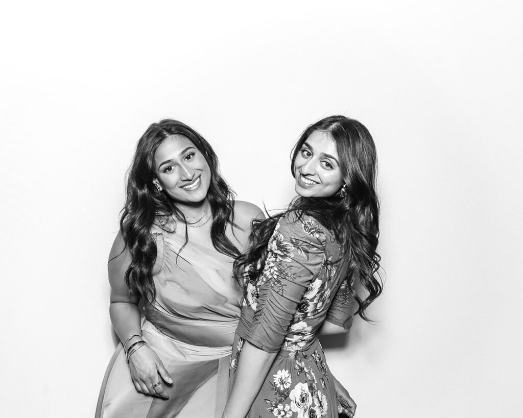 Two friends smiling and posing side by side in a Kardashian-style black and white wedding photo booth, dressed in elegant floral and draped gowns