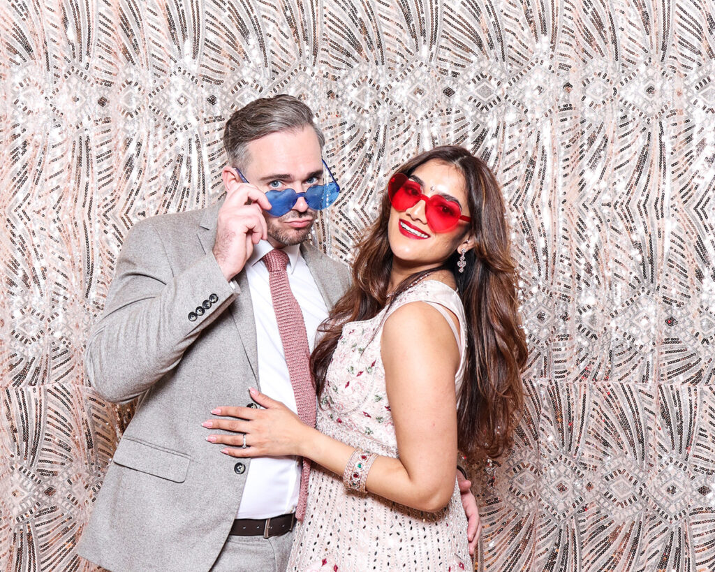Modern couple in playful photo booth moment at luxury Asian wedding with sequin embellished backdrop