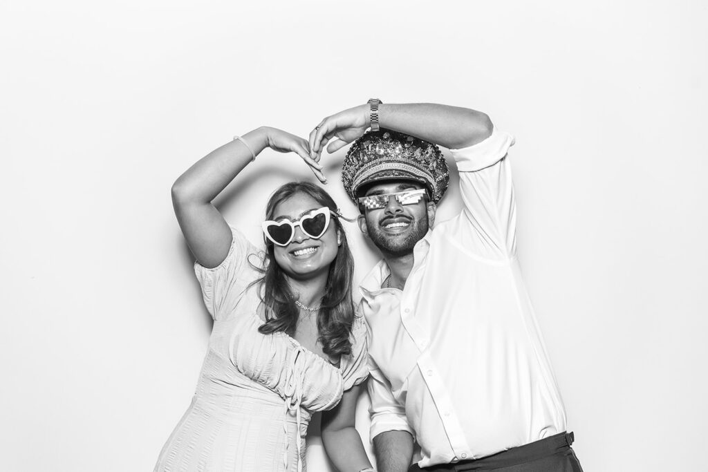 Couple forming a heart shape with their arms, wearing statement glasses and crowns, captured in a Kardashian-style black and white photo booth moment
