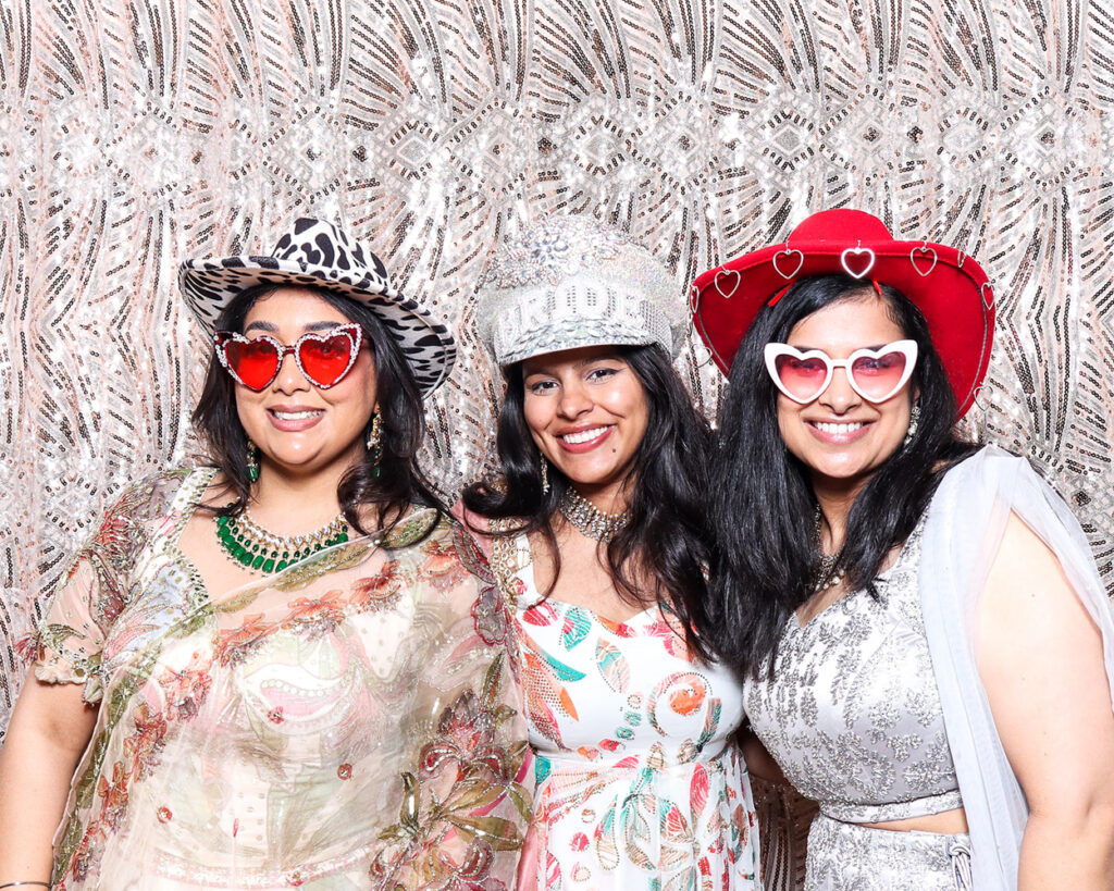 Three wedding guests in colourful South Asian outfits and fun heart-shaped sunglasses, posing with playful hats against a sparkly sequin photo booth backdrop