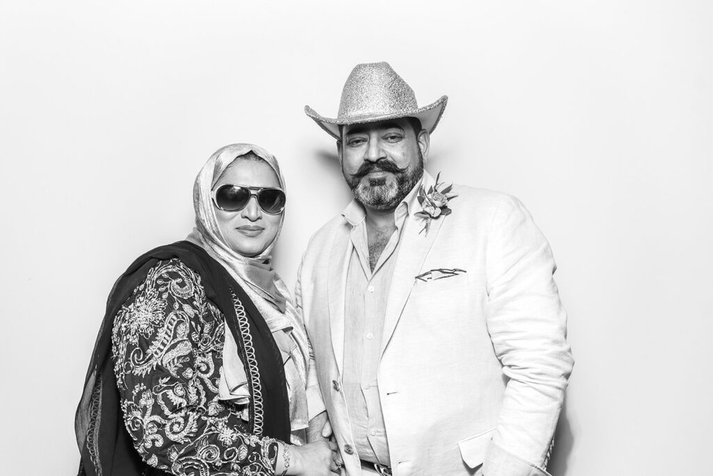 Stylish couple posing confidently in a black-and-white Kardashian-style photo booth shot—one in a shimmering headscarf and sunglasses, the other in a cowboy hat and light suit with a boutonnière
