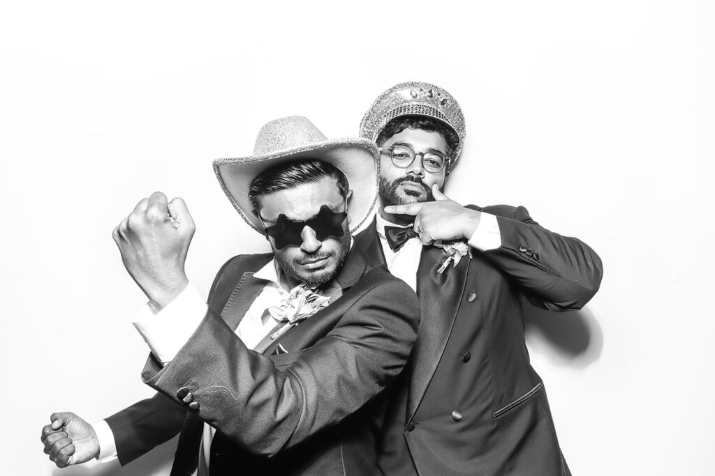 Wedding guests striking bold poses in tuxedos with fun props inside a Kardashian-style black and white photo booth setup