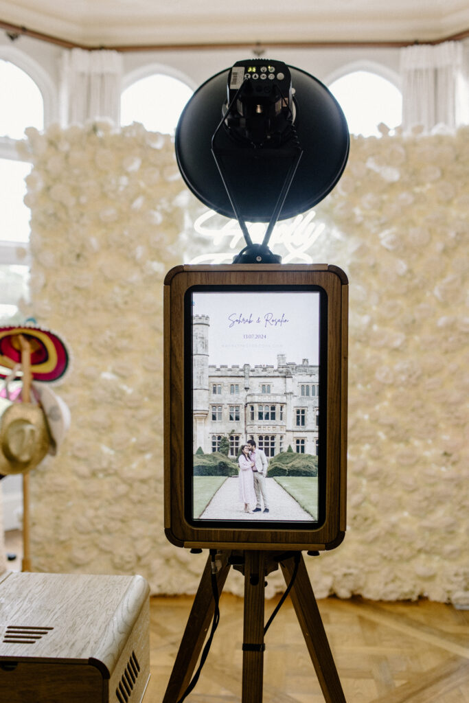 The back of the Icon photo booth setup for an asian wedding celebration, at Farleigh House with a white roses floral backdrop and a neon sign over