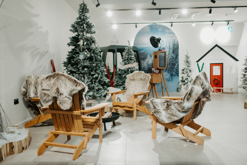 Apres ski themed Christmas photo booth lounge at Cotswolds Designer Outlet, with wooden chairs, faux fur throws, snowy trees and a wooden photo booth against a winter mountain backdrop.