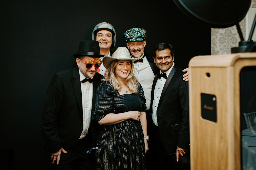 Team of guests in formal wear posing together for a picture at a black backdrop corporate Christmas celebration.