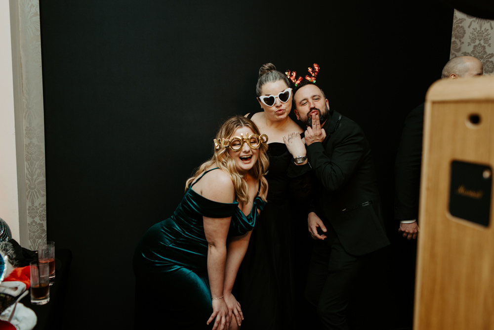 two women and one man in black-tie outfits posing playfully for a photo booth picture, one wearing heart-shaped glasses.