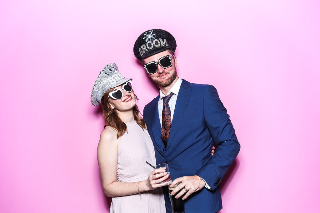 couple posing against a bright bubble pink backdrop for a Wedding Photo Booth with Bride and Groom Hat Props during a wedding party entertainment