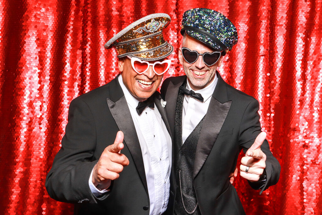 2 men posing with party props against a red bright backdrop for a Wedding Photo Booth with Props during a wedding party entertainment